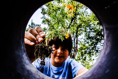 Low angle portrait of smiling boy holding flowering plant against trees