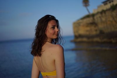 Rear view of young woman looking away while standing against sea during sunset