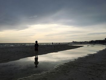 Silhouette boy standing at beach against cloudy sky