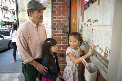 Girl showing menu to grandfather and sister while standing against restaurant