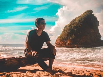 Man sitting on rock at beach against sky