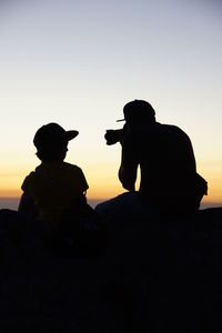 Silhouette men sitting on rock against sky during sunset