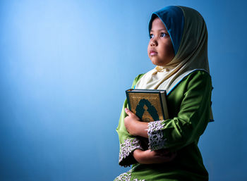 Girl wearing hijab holding koran while sitting against blue background