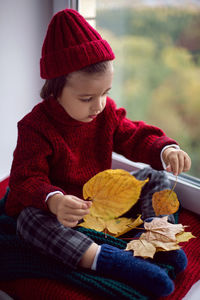 Boy child in red knitted sweater and hat sitting on the window in autumn