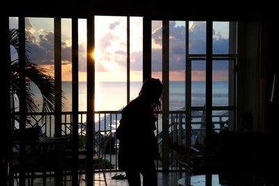 Woman standing by window against cloudy sky during sunset