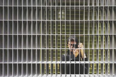 Man photographing while standing behind metallic fence