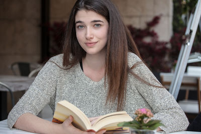 Portrait of smiling young woman holding book at sidewalk cafe