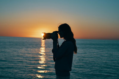 Silhouette man photographing sea against sky during sunset