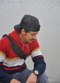 A young guy wearing cap backwards, looking down while sitting next to the lake