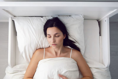 Woman in a white t-shirt is lying on the bed and a pillow is under the blanket in the room