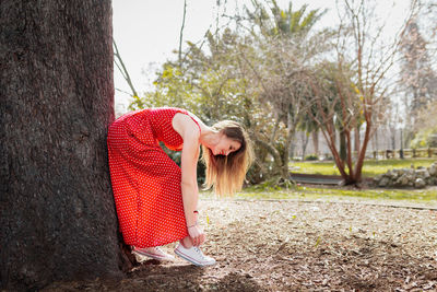 Woman in red dress tying shoelace against tree trunk