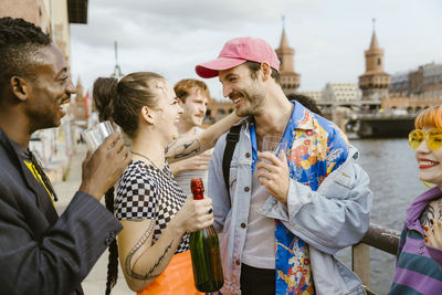 Smiling gay man talking with non-binary friend holding champagne bottle on promenade in city