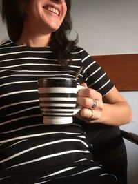 Midsection of happy woman holding striped coffee cup while sitting against wall