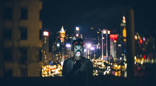 Man wearing mask while using mobile phone against cityscape at night