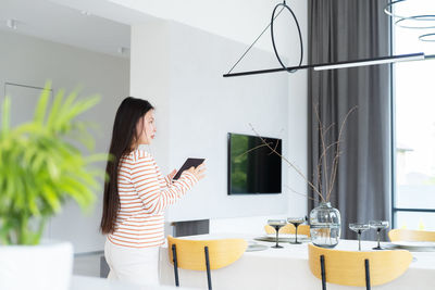 Asian woman controlling smart home functions through tablet at modern home, looking at ceiling lamp