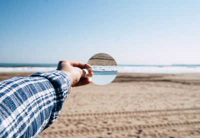 Cropped hand holding mirror with reflection against sea