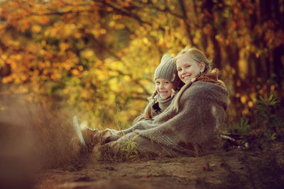 Lovely sisters are sitting, wrapped in a warm blanket, in an autumn park among the golden trees 