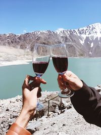 Cropped hands toasting red wineglasses against lake