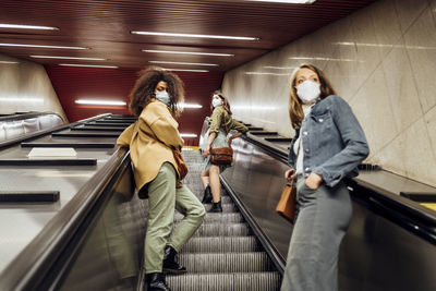 Friends with protective face mask standing at social distance on escalator