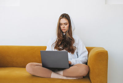 Young woman in white hoodie beautiful girl freelancer student using laptop sitting on yellow couch 