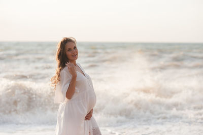 Beautiful laughing pregnant woman wear white dress posing over sea at background closeup.