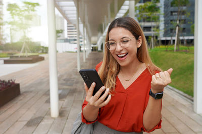 Beautiful successful brazilian female excited raised hand rejoicing with smartphone outside. 