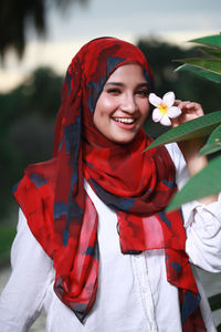 Portrait of cheerful young woman wearing hijab while holding flower by plant