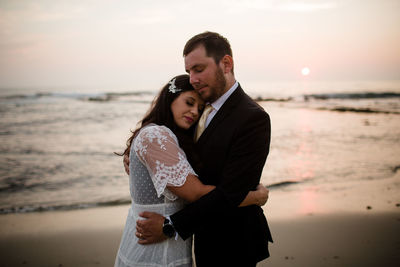 Couple standing at beach against sky during sunset