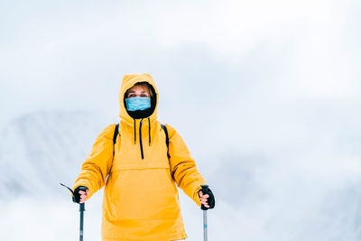 Unrecognizable female hiker in yellow outerwear and protective mask looking at camera while standing with trekking poles in snowy mountains