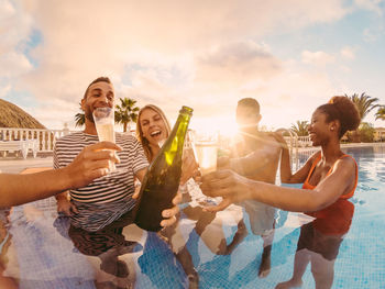 Friends toasting beer bottles and glasses in swimming pool against sky during sunset