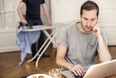 Gay man using laptop at table with partner ironing in background