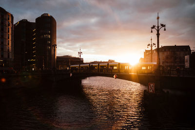 Bridge over river by buildings against sky during sunset