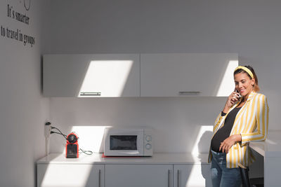 Young pregnant female in trendy striped jacket and jeans talking on smartphone while standing in modern kitchen with white furniture and microwave oven