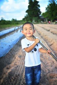 Portrait of smiling boy with rake standing on farm against sky