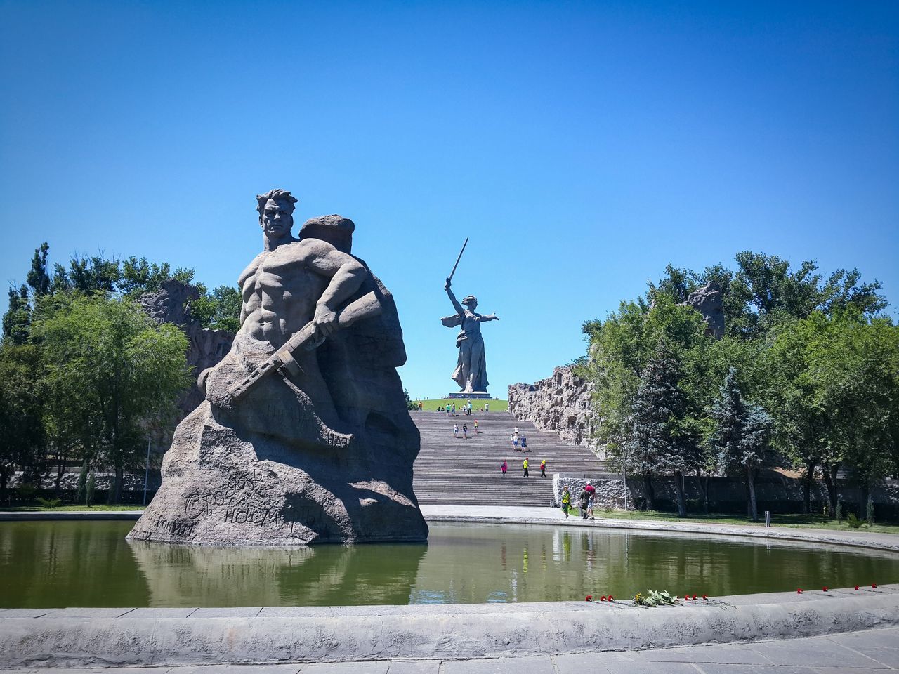 clear sky, water, blue, copy space, tree, statue, men, sculpture, human representation, nature, leisure activity, tranquility, lifestyles, waterfront, day, rock - object, tranquil scene, sunlight