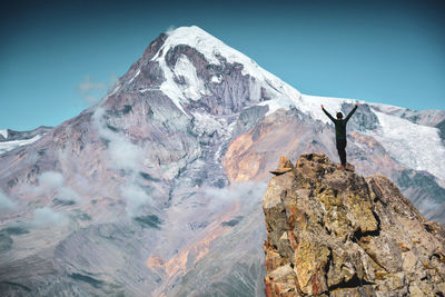 Rear view of young man standing on rock formation with arms outstretched against mountain and sky