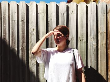 Woman looking away while shielding eyed against wooden fence