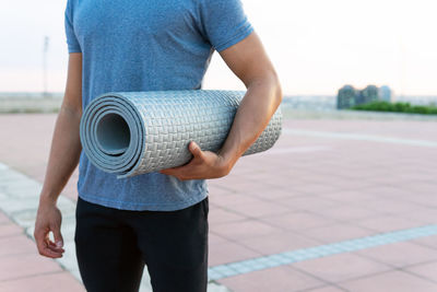 Cropped unrecognizable male in sportswear standing with twisted yoga mat on street and looking away