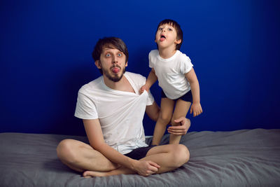 Boy child with his father in white t shirts sitting at home on a bed against a blue wall