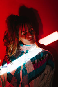 Young female in old fashioned outfit looking into distance while standing near wall under ray of light in dark studio