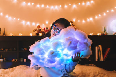 Woman holding illuminated cloud while sitting on bed at home