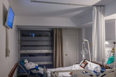 Side view of elderly female patient in medical mask lying on bed in hospital ward with visitor male wearing protective uniform sitting in armchair