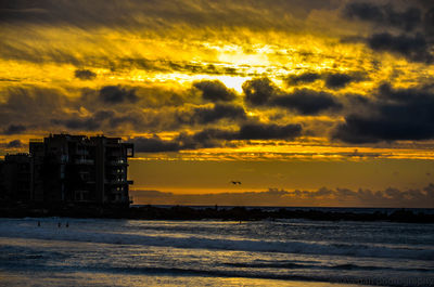 Silhouette buildings by sea against sky during sunset