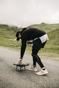 Side view of man holding quadcopter while standing on road against sky