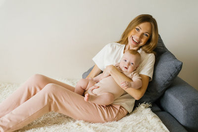 Smiling mother is sitting on the sofa and holding her baby son in her arms and smiling at him