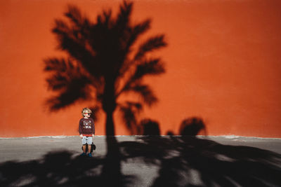 Boy standing against a red wall with the shadow of a palm tree