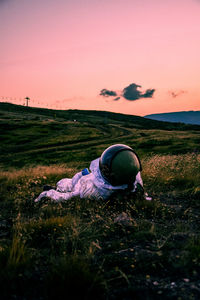 Rear view of an astronaut sitting on grassy field against sky during sunset