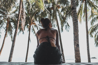 Rear view of shirtless woman sitting against palm trees