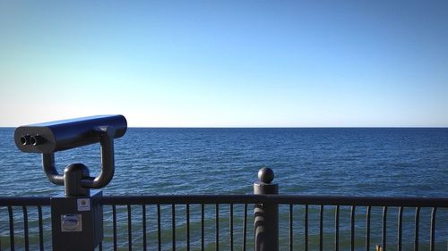 Metal railing by sea against clear blue sky