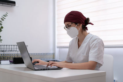 Concentrated female doctor in medical mask and uniform sitting at table and browsing laptop while working in modern workspace